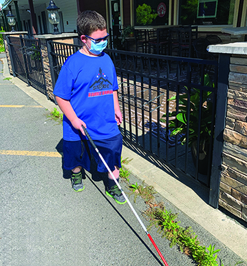 A White teenage boy wearing a mask uses his cane to shoreline a curb.” width=“460