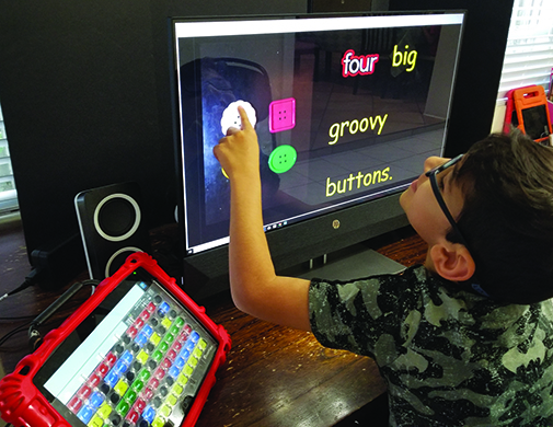 A Multiracial school-age boy points to a white button on a black screen showing four buttons and the words 