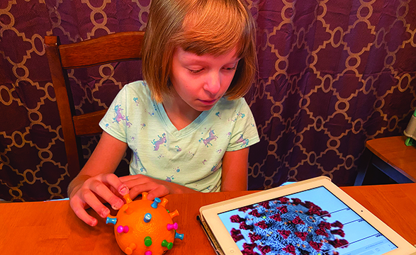 A White school-age girl examines a tactile model of COVID-19 made from push-pins on an orange. She compares the tactual model to an enlarged image of COVID-19 on her iPad screen.