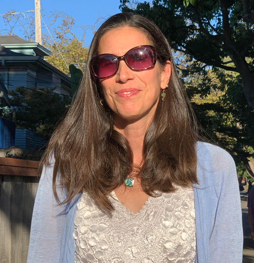 Rachel Longan. Woman with long brown hair. She is standing outside, wearing sunglasses, a light blue cardigan, and white blouse.