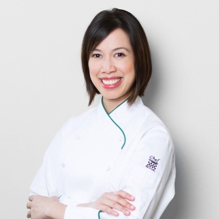 Christine Ha has dark neck-length hair. She wears a white chef's jacket. She is smiling with arms crossed in front of her. 