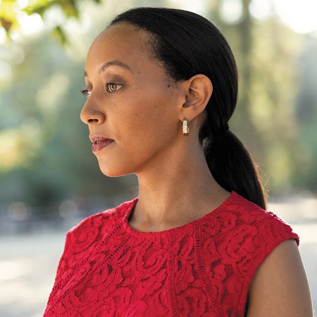 Haben Girma stands outdoor on a sunny day. She is viewed in three-quarter profile and is looking off to her right,, her face is pensive. Her long dark hair is pulled back in ponytail fashion and golden hoops adorn her ears. Haben is wearing a sleeveless rose-colored dress with raised patterns.
