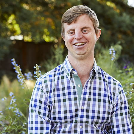 Dr. Hoby Wedler standing outside in a garden, wearing a checked shirt with collar.