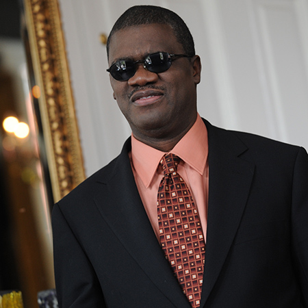 Marcus Roberts is standing in an elegant hall. Over his right shoulder is a large wall mirror in a gold gilt frame. Marcus is a black man with short cropped hair. He's wearing sunglasses, a peach color dress shirt with a patterned tie under a charcoal grey two button blazer. 