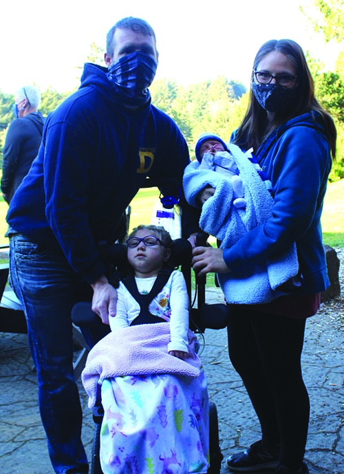 A White preschool deafblind girl is sitting in her wheelchair next to her family. Her mom and dad are wearing masks. Her mom is holding her newborn brother. They are outside.