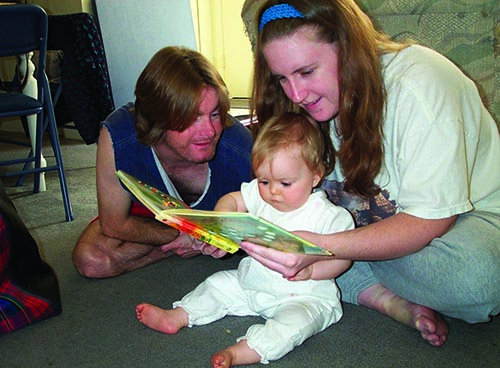 A White infant girl shares a board book with her white mom and dad. They all sit on the floor and mom holds the book
