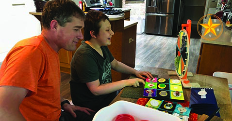 A White teenager sits at a kitchen table smiling with his hands resting on a game that has been adapted using tactile symbols. His father sits beside him and his instructor joins them online via an iPad resting on the table.