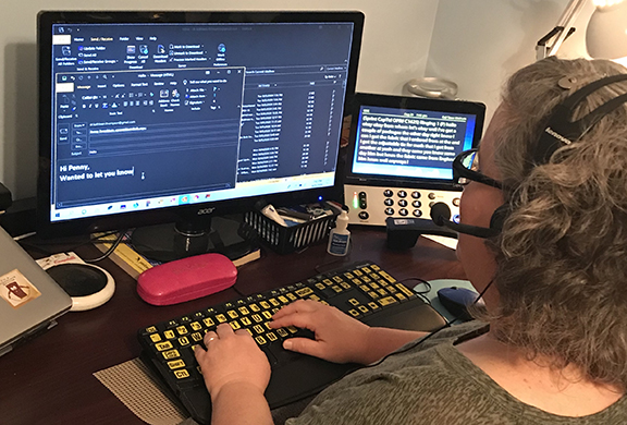 View from behind of a woman wearing glasses and a headset sitting at a computer with a large print keyboard and high contrast screen