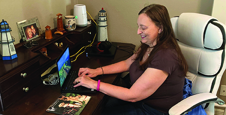 A smiling woman with light skin and long dark hair sits in an office chair at a desk with her hands on a laptop keyboard